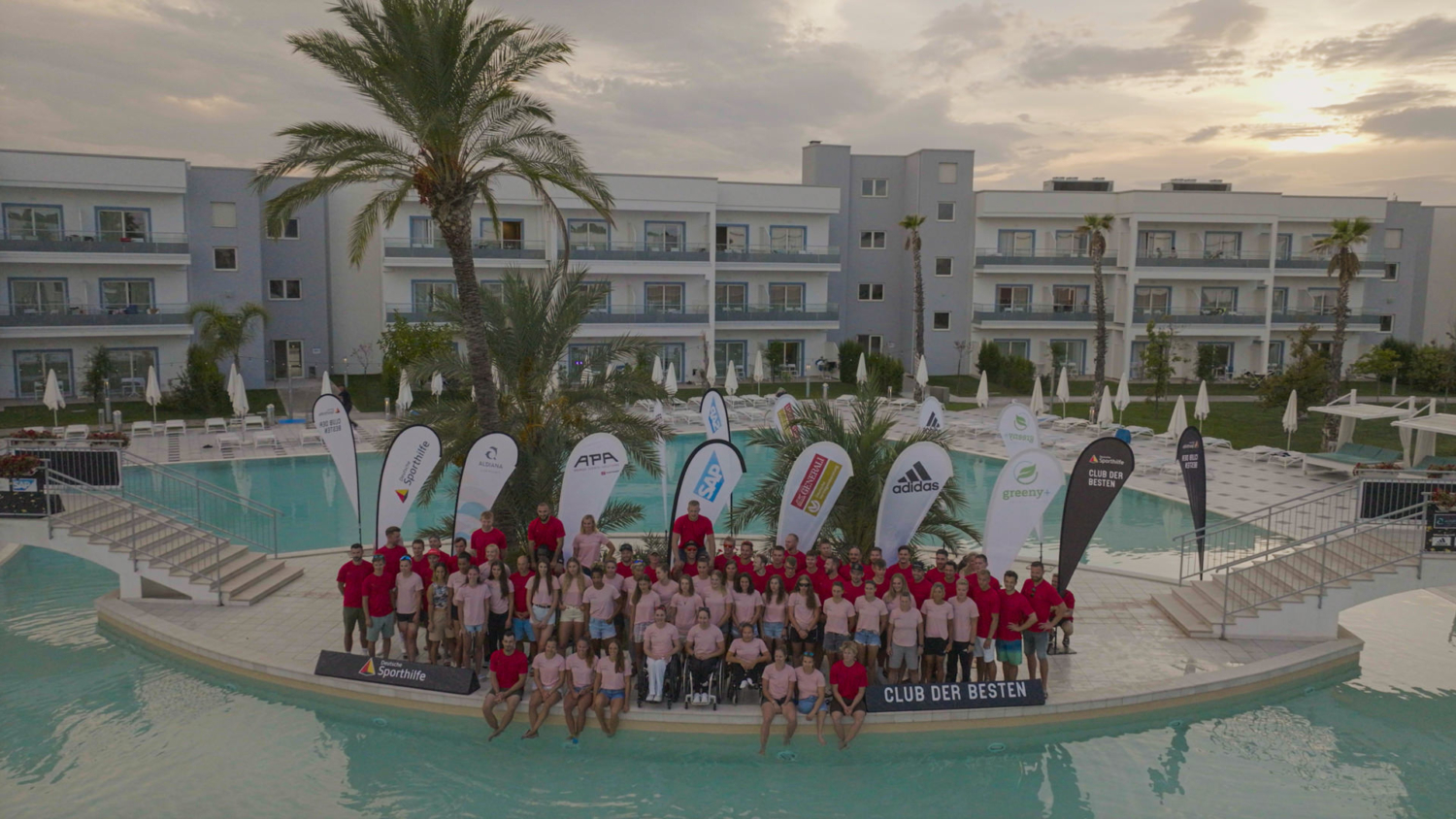 Eine große Gruppe versammelt sich für ein Gruppenfoto an einem Pool, umgeben von Palmen und Werbebannern bekannter Marken – ein Event in exklusiver Atmosphäre.