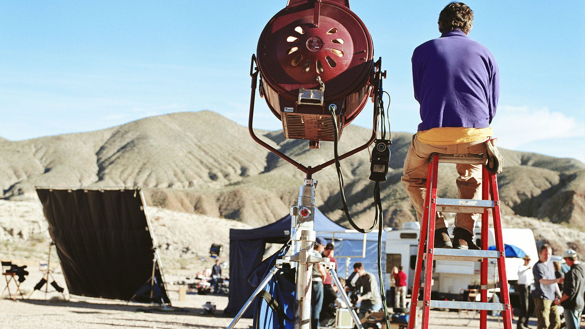 Filmset mit großer Lichtquelle, Person auf Leiter, Berge im Hintergrund – Aufbauphase am Außendreh