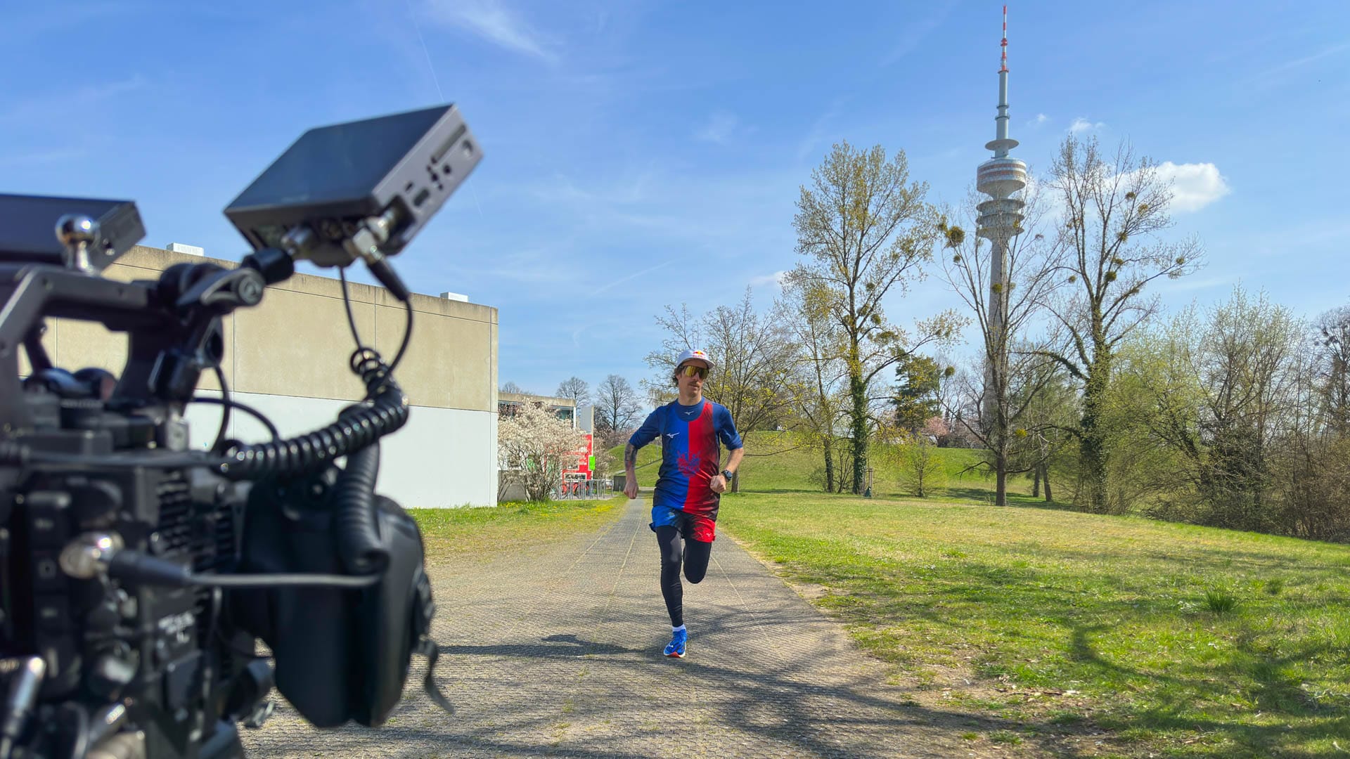 Kamera filmt Flo Neuschwander beim Laufen im Grünen mit dem Olympiaturm im Hintergrund