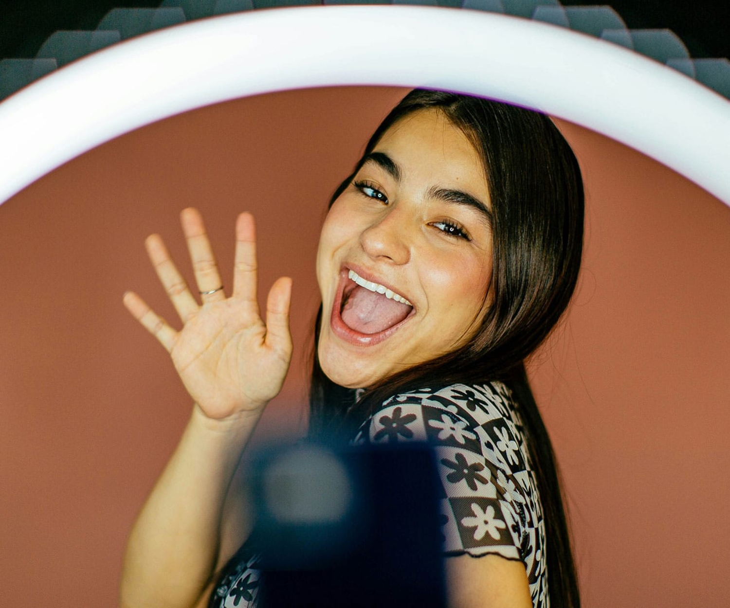 Junge Frau winkt fröhlich in die Kamera beim Erstellen von User-Generated Content im Unternehmensvideo mit Ringlicht