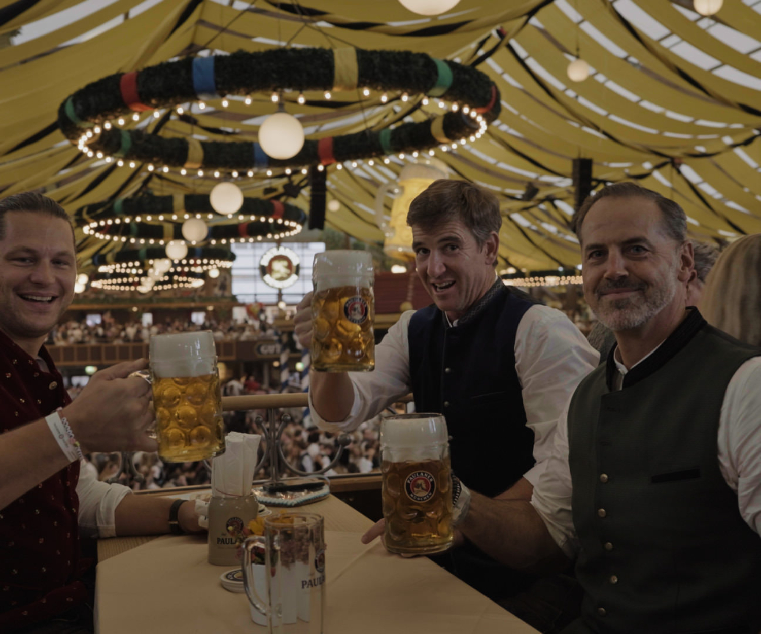 Markus Kuhn, Eli Manning und Shaun O’Hara stoßen mit Maßkrügen im Paulaner Festzelt an